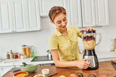 Mutfakta lezzetli vejetaryen içeceği yaparken blender 'da taze meyve öğüten gülümseyen kadın.