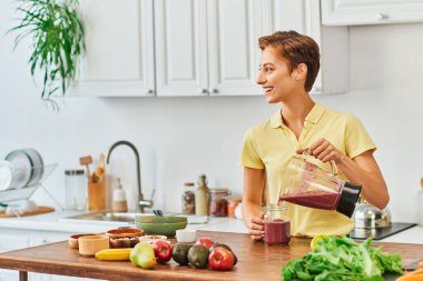 Neşeli kadın blenderdan mason kavanozuna vejetaryen smoothie dolduruyor, bitki temelli diyet konsepti.
