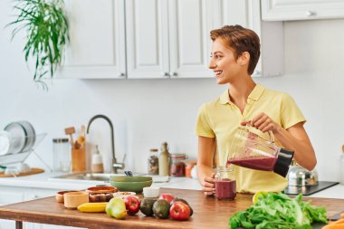 cheerful woman pouring vegetarian smoothie from blender into mason jar, plant-based diet concept clipart