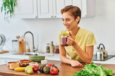 Gülümseyen vejetaryen kadın mutfaktaki samanla mason kavanozundan lezzetli bir içecek içiyor.