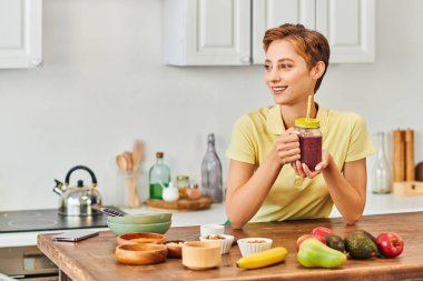 Mutfaktaki taze meyvelerin yanında bitki bazlı smoothie ile mason kavanozu tutan gülümseyen kadın.