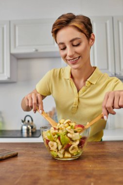 Mutfakta taze meyve salatasıyla ahşap spatulaları karıştıran neşeli kadın. Lezzetli vejetaryen tarifi.