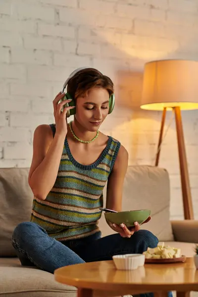 vegetarian woman with salad bowl listening music in headphones near tofu cheese in living room