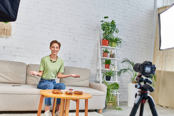 smiling woman presenting vegetarian meal during video blog on plant-based diets in green living room