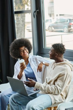 joyful friends in casual outfits with coffee cup and laptop smiling at each other, business concept clipart