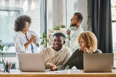 two cheerful young people working on laptop with their friends on backdrop, business concept clipart