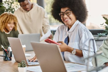 Gözlüklü neşeli genç bayan kameraya gülümsüyor. Arkadaşları projede çalışıyor.