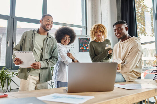 four jolly friends looking at laptop working on project with notes and coffee cup in hands, business