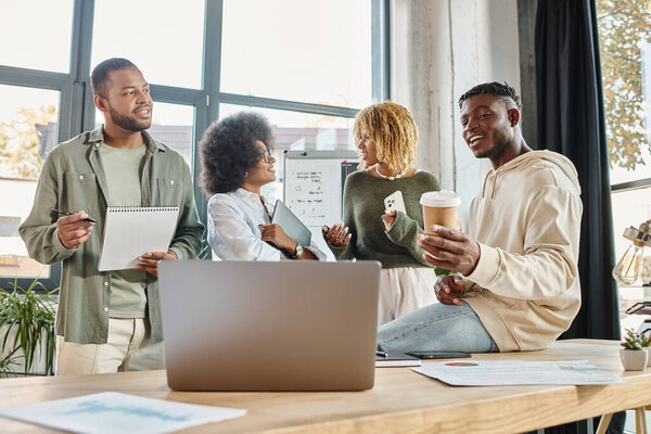 joyful friends discussing project other with laptops and notes in hands, coffee in hand, business