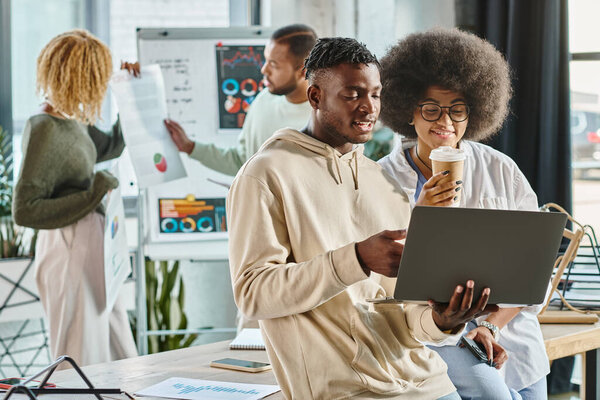 cheerful man and woman looking at laptop while their friend discussing graphs, coworking concept