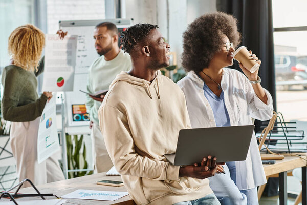 joyous team members with coffee and laptop in hands with their friends on backdrop, working process
