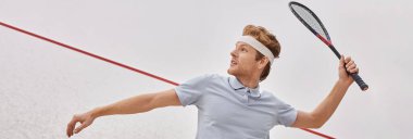 Spor giyim ve saç bantlı kızıl saçlı adam raket tutuyor ve sarayın içinde squash oynuyor.