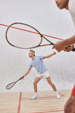 focus on redhead man in sportswear playing with african american friend inside of squash court clipart