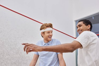 Raketlerle işaret eden mutlu ırklar arası oyuncular ve squash sahasının içine bakan arkadaşlar.