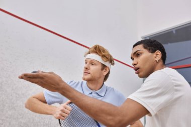 Genç Afrikalı Amerikalı adam squash topu tutuyor ve sarayın içindeki arkadaşına bir şey gösteriyor.