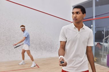 Genç Afrikalı Amerikalı adam squash topu tutuyor ve sarayın içinde kızıl saçlı arkadaşıyla oynuyor.