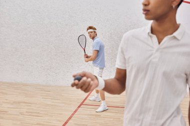 redhead man holding racquet near african american man with squash ball playing in court clipart