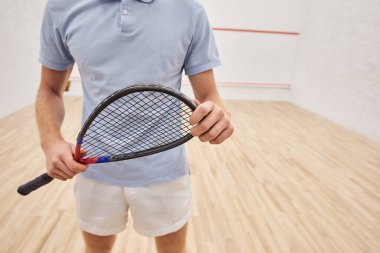Spor kıyafetleri içinde raket tutan ve iç sahada dikilen bir adamın görüntüsü.