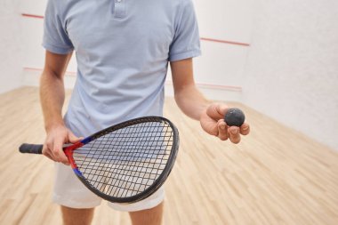 Spor kıyafetleri içinde elinde duvar tenisi topu, raket ve mahkeme salonunda dikilen bir adamın görüntüsü.