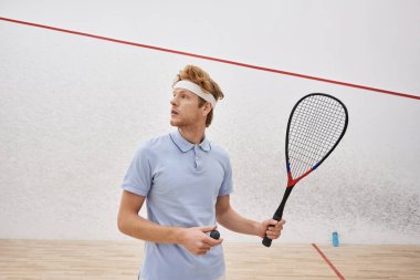 Spor kıyafetli kızıl saçlı adam iç sahada dururken elinde squash topu ve raket tutuyor.