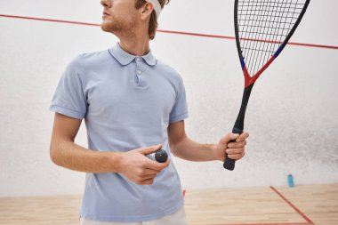 Spor giyim kuşağında, elinde squash topu ve raket tutan bir adamın mahkemenin içinde dikilirken çekilmiş görüntüsü.