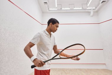 Aktif görevde olan Afro-Amerikan bir adam mahkeme salonunda oynarken squash topu ve raket tutuyor.