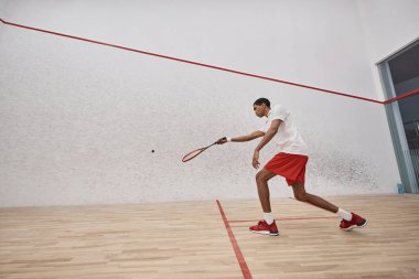 Kırmızı şortlu aktif Afro-Amerikan sporcusu sarayın içinde squash oynarken raket tutuyor.