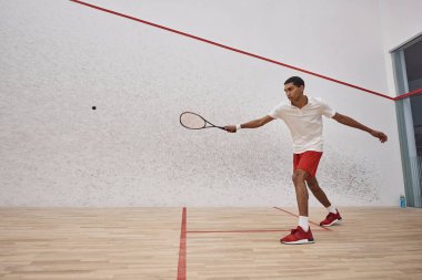 Kırmızı şortlu aktif Afro-Amerikan sporcusu raket tutuyor ve mahkemede squash oynuyor.
