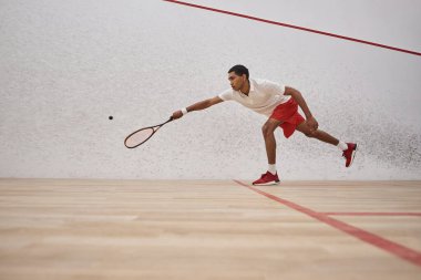 Kırmızı şortlu Afro-Amerikan sporcusu raket tutuyor ve sarayın içinde squash oynuyor.