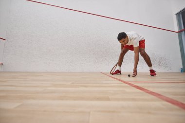 Kırmızı şortlu Afro-Amerikan bir adam raket tutuyor ve sarayın içinden squash topu topluyor.