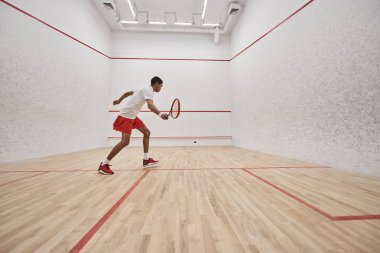 Spor şortlu Afro-Amerikan sporcu sarayın içinde squash oynarken raket tutuyor.