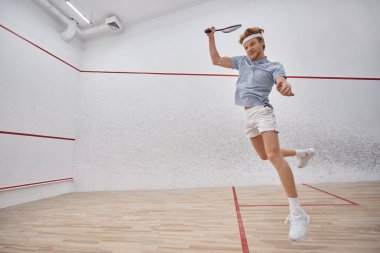 motion shot, energetic sportsman holding racquet and jumping while playing squash inside of court clipart