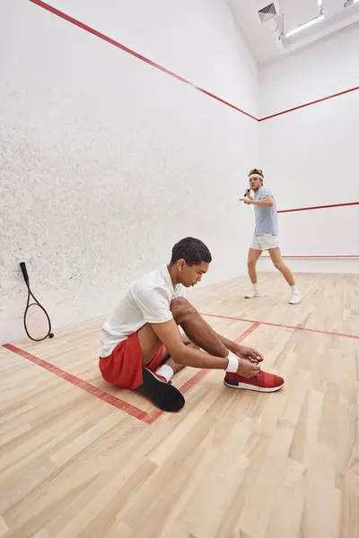 Stock image african american man tying shoelaces while sitting near redhead friend inside of squash court