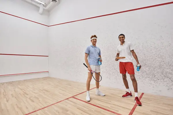 stock image cheerful interracial sportsmen with squash racquets and bottles with water standing inside of court