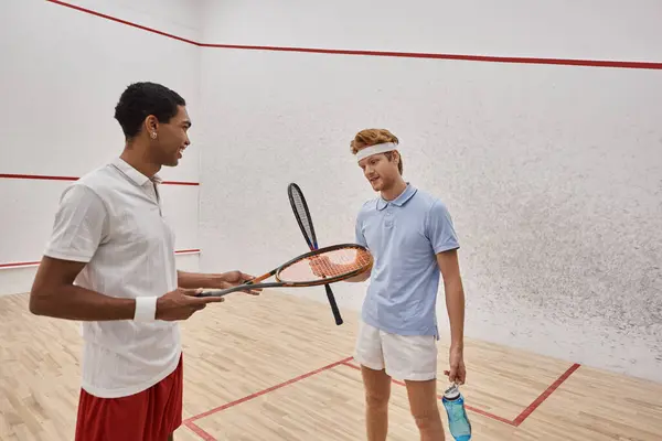 stock image joyful interracial male friends with squash racquets and bottles with water chatting  in court