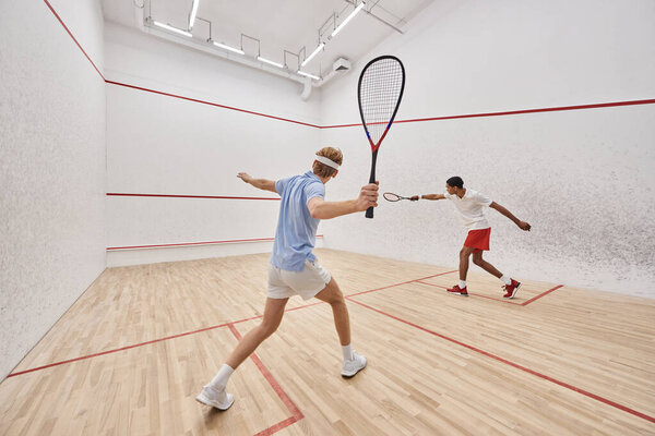 two multicultural players in sportswear playing squash together inside of court, active lifestyle