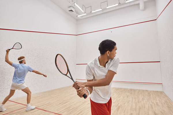 interracial friends in sportswear playing squash together inside of court, active lifestyle