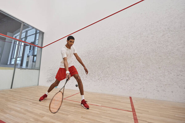 young african american man in red sporty shorts holding racquet while playing squash, motion shot