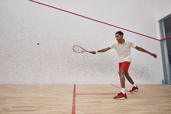 active african american sportsman in red shorts holding racquet and playing squash inside of court