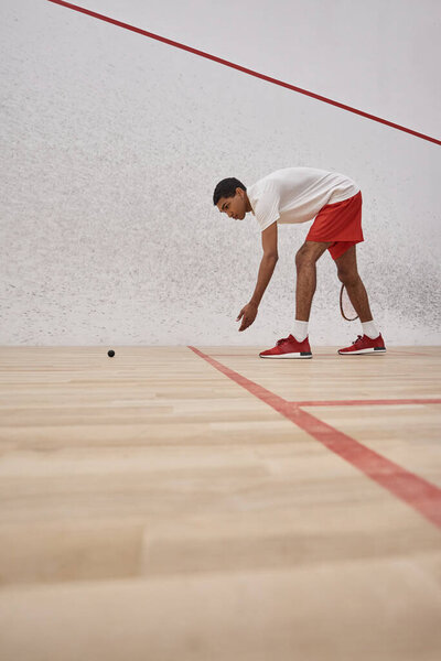 african american man in red sporty shorts holding racquet and picking up squash ball inside of court