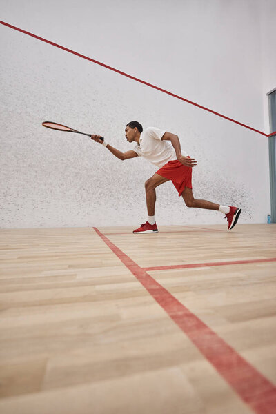energetic african american sportsman in shorts holding racquet and playing squash inside of court