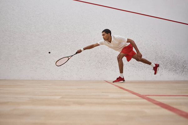 african american sportsman in red shorts holding racquet and playing squash inside of court, sport