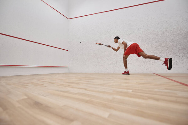african american sporty man in red shorts holding racquet while playing squash inside of court