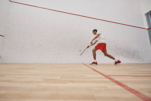 african american young man in red shorts holding racquet while playing squash inside of court