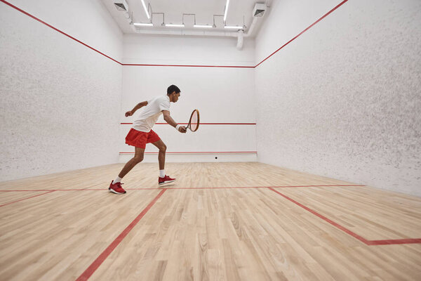 african american sportsman in sports shorts holding racquet while playing squash inside of court