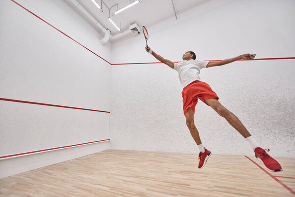 motion shot, energetic african american sportsman with racquet jumping and playing squash in court