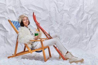 happy asian model in winter clothes sitting in deck chair with cocktail and skis in snowy studio clipart