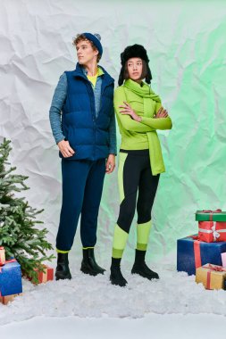 Sıcak giyimli çok ırklı çift hediye kutularının yanında ve Noel ağacı karlı stüdyoda, bayram mevsiminde.