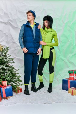 Sıcak giyimli çok ırklı çift hediye kutularının yanında ve Noel ağacı karlı stüdyoda, bayram mevsiminde.