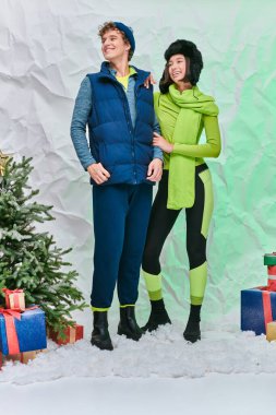 Hediyelerin yanında sıcak giysiler içinde neşeli ırklar arası modeller ve stüdyoda kar üzerinde Noel ağacı.